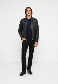 Black leather jacket with a smooth texture, featuring a high collar and front zipper. Worn over a navy sweater and fitted black pants.