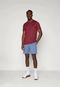 Bordo sporta krekls ar īsām piedurknēm; gaiši zili šorti ar logo; balti sporta apavi; neitrāla fonā. Uzsvars uz ikdienišķas sporta apģērbu dizainu.