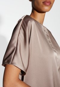 Satinbluse in warmem Taupe mit kurzen, sanft drapierten Ärmeln und rundem Ausschnitt, mit einer glatten, glänzenden Textur.