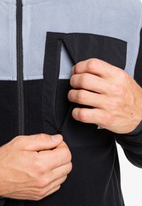 Futrzana kurtka z czarnymi, szarymi i niebieskimi panelami, z zamykaną kieszenią. Zbliżenie na ręce zapinające kieszeń. Miękka tekstura.