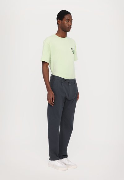 Lys grøn t-skjorte med kaktusgrafikk, kombinert med mørkegrå bukser og hvite sneakers. Enkel design, avslappet passform, stoffets tekstur ikke synlig.