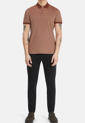 Polo marron avec col et patte à trois boutons, avec bordure marron foncé au col et aux manches. Porté avec un pantalon noir et des chaussures.