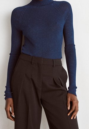 Vrouw draagt een aansluitende donkerblauwe geribbelde coltrui en hoge taille zwarte geplooide broek tegen een effen achtergrond.