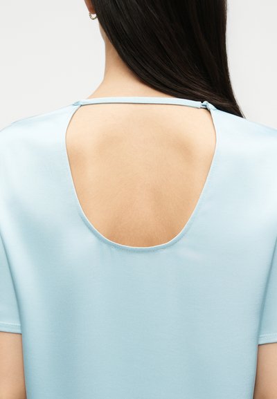 Γυναίκα με μακριά σκουρόχρωμα μαλλιά φοράει ανοιχτό μπλε τοπ με μεγάλο οβάλ άνοιγμα στην πλάτη, το οποίο στερεώνεται με λεπτή λουρίδα στο λαιμό.