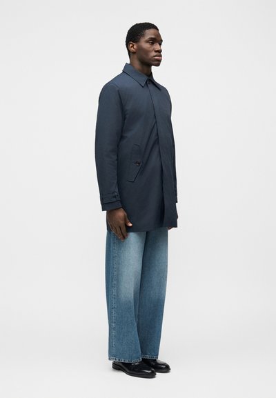 Μπλε σκούρο παλτό με κλασικό γιακά, δύο μπροστινές τσέπες και κουμπιά· συνδυασμένο με ανοιχτό μπλε τζιν φαρδιάς γραμμής και μαύρα παπούτσια.