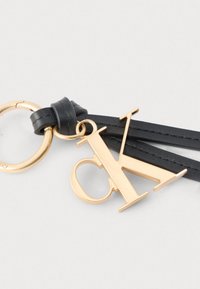 Χρυσός μεταλλικός γοητευτικός λογότυπος CK προσαρτημένος σε μαύρα δερμάτινα λουράκια και χρυσό κρίκο κλειδιών σε ανοιχτό γκρι φόντο.