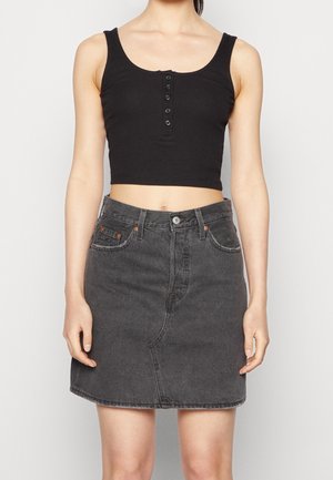 Kobieta ubrana w czarny, bezrękawnik z guzikami i wysoką, wyblakłą czarną spódnicę z denimu, stojąca na jednolitym tle