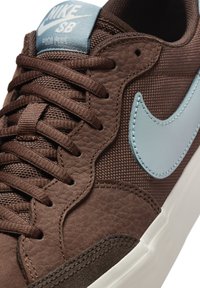 Brun Nike SB Pogo Plus sneaker med tekstureret brun læder, mesh-detaljer, blå swoosh-accent og hvid gummisål. Snørebåndene er brune.
