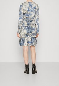 Femme portant une robe froncée fleurie bleue et crème avec des manches longues et des bottines noires, se tenant de dos contre un fond uni.