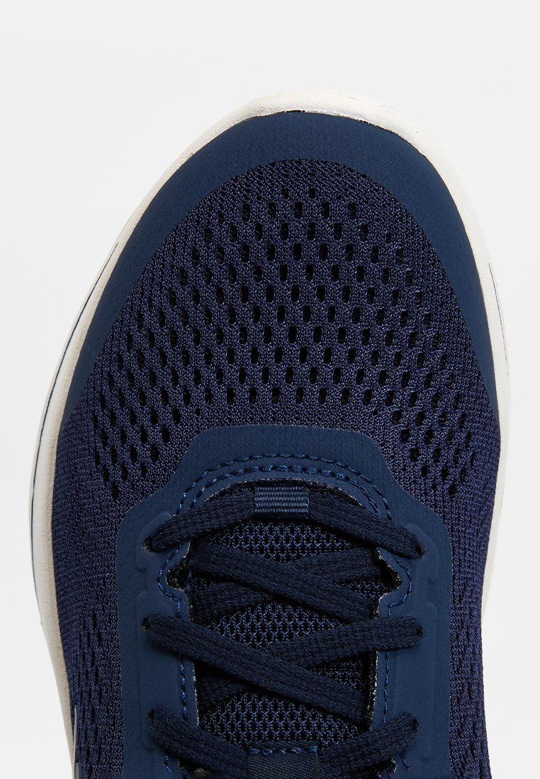 Zapatilla deportiva azul marino con parte superior de malla, puntera reforzada y cordones tonales. Presenta una suela de goma blanca y un talón estructurado para soporte.