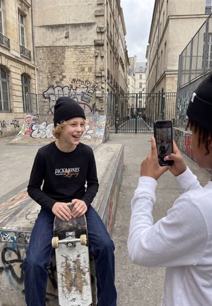 Un adolescent souriant est assis sur un skateboard dans un skatepark couvert de graffitis, tandis qu’une autre personne le photographie avec un téléphone.