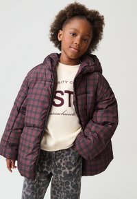 Doudoune à carreaux bordeaux avec capuche, présentant une texture matelassée et des poches latérales. Associée à un sweat crème et un pantalon imprimé léopard.