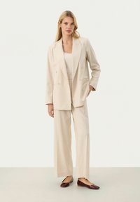 Beige skräddarsydd kostym med en dubbelfransad kavaj och vida byxor, med en slät yta och minimala detaljer. Kombinerad med vinröda lägenheter.