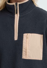 Fleece-genser med høy krage, mørk marineblå farge, kontrasterende beige glidelås og lomme, strukturert stoff og kvart-glidelås foran.