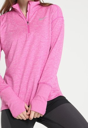 Kobieta ubrana w różowy top Nike z długim rękawem, zamkiem do połowy i otworami na kciuki, w połączeniu z czarnymi legginsami o fakturze, na jednolitym tle.