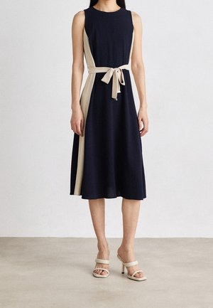 Femme portant une robe midi sans manches bleu marine avec des panneaux latéraux beige et une ceinture nouée, associée à des sandales à talons hauts beiges.