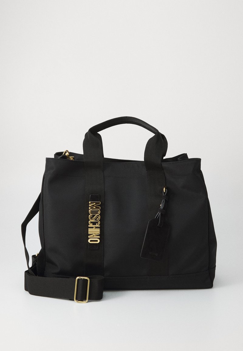 Saco tote preto da Moschino com alças duplas, alça de ombro ajustável, lettering em dourado e etiqueta de bagagem preta presa em fundo liso.