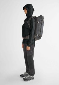 Veste noire à capuche avec zip, pantalons gris foncé arborant des patchs texturés, et un sac à dos noir. Équipé de sangles réglables et de boucles.