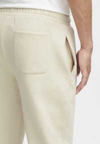Beige színű, textúrált anyagú melegítőnadrág, gumis derékkal, oldalsó zsebekkel és hátsó zsebbel. Letisztult, minimalista dizájn.