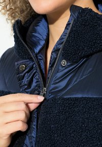 Granatowa kurtka z miękkim, teksturowanym kołnierzem z polarowego materiału. Posiada centralny zamek błyskawiczny oraz napy, prezentując projekt z mieszanych materiałów.