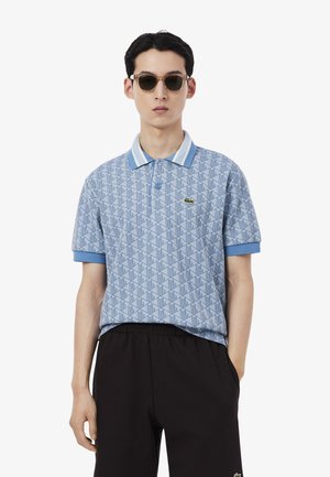 Homme portant un polo bleu à motifs, un pantalon noir et des lunettes de soleil, debout avec une main dans la poche, sur fond blanc.