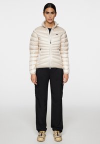 Beżowa kurtka ocieplana z zamkiem błyskawicznym, wysokim kołnierzem i detalem logo, zestawiona z czarnymi spodniami cargo i beżowym obuwiem. Gładka tekstura.