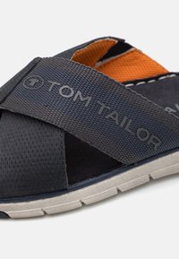 Navyblaue Sandale mit einem strukturierten Kreuzriemen, der ein schwarzes Logo und ein-orangefarbenes Innenfutter aufweist. Gummisohle mit einem hellgrauen Profil.