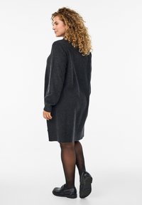 Ciemnoszara dziana sukienka z długimi rękawami, prążkowanymi mankietami i dołem, połączona z czarnymi butami i półprzezroczystymi czarnymi rajstopami. Modelka jest ustawiona z boku.