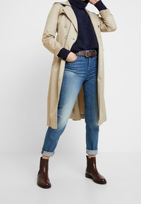 Beige trenchcoat över en mörk tröja, tillsammans med blå jeans med uppvikta ben och bruna anklarstövlar. Innehåller knappar och ett bälte.