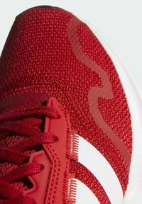 Czerwony but sportowy z teksturowaną cholewką z dzianiny, białymi akcentami i czarną gumową podeszwą. Posiada sznurowadła oraz wyraźny wzór na boku.