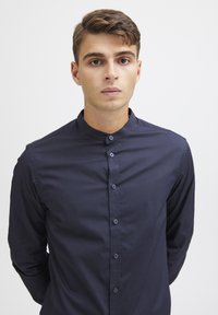 Camisa de botones azul marino con cuello de banda, mangas largas y una textura suave. Cuenta con botones frontales y un diseño ajustado.