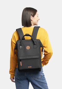 Zaino in nylon nero con spallacci doppi, tasca frontale con zip e manico superiore. Presenta un logo circolare e dettagli in pelle.