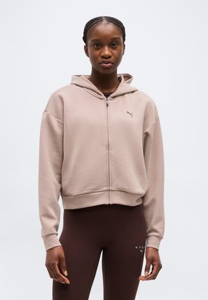 Woman with braided hair wearing a beige zip-up hoodie and dark brown leggings standing against a plain white background.