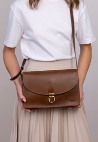 Hnědá kožená křížová taška s zakřiveným klopou, zlatou kovovou sponou a detailem stehu. Hladký povrch a obdélníkový tvar.