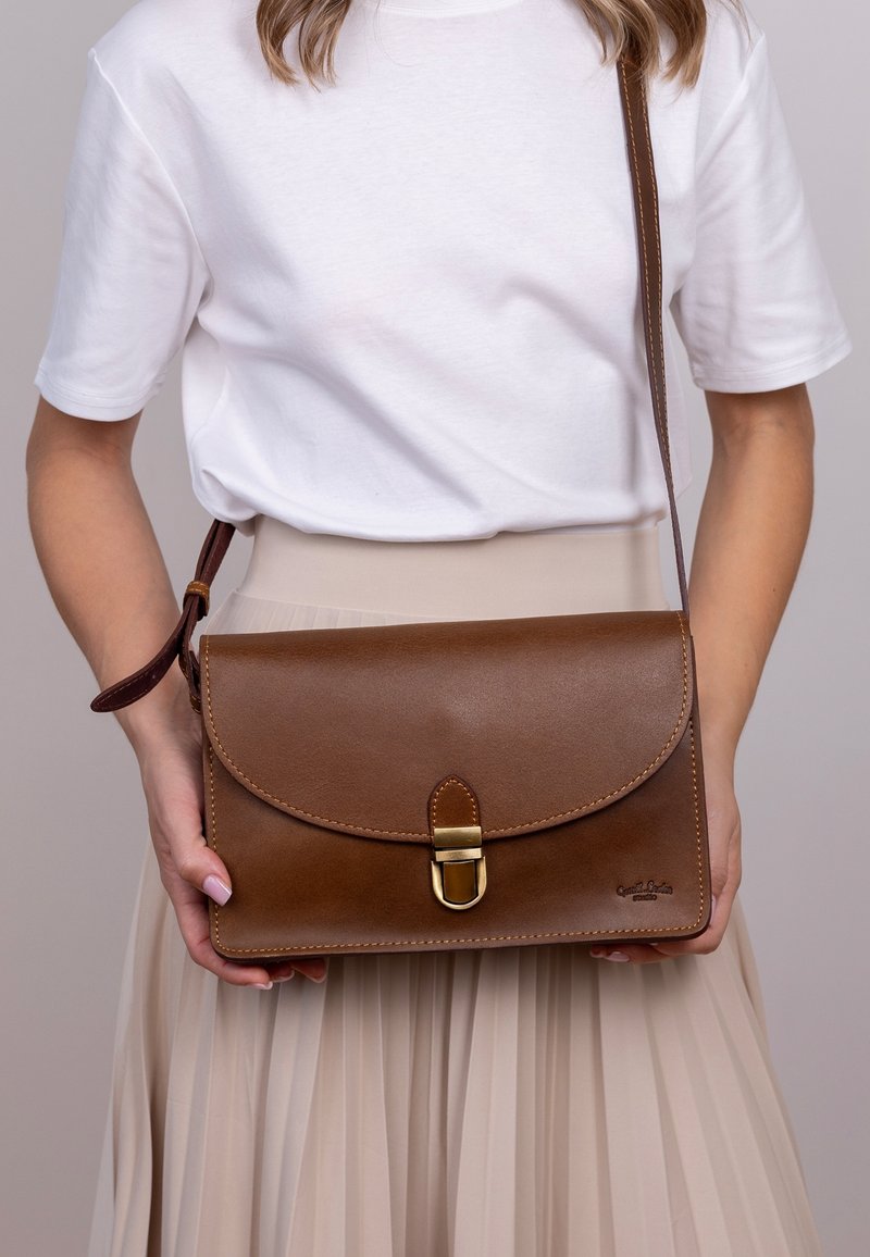 Hnědá kožená křížová taška s zakřiveným klopou, zlatou kovovou sponou a detailem stehu. Hladký povrch a obdélníkový tvar.