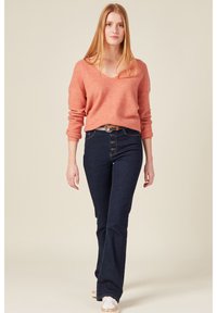 Helles rosa, strukturiertes Sweatshirt mit V-Ausschnitt, kombiniert mit dunkelblauen, weiten Jeans mit Knopfverschluss und einem braunen Gürtel. Weiße Sneakers runden den Look ab.
