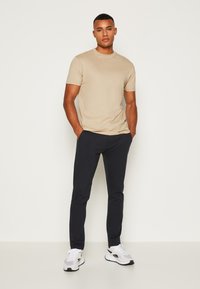 Beige, rövid ujjú póló sötét tengerészkék, tapered nadrággal és fehér sportcipővel párosítva. Az anyag simának tűnik, és fitted fazonú.