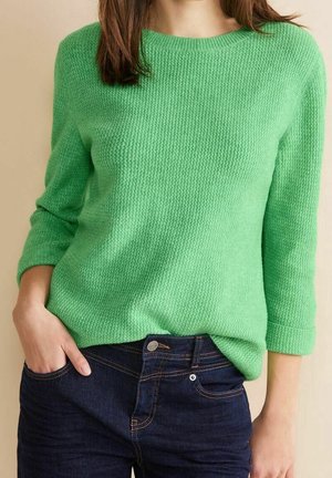 Frau trägt einen strukturierten grünen Pullover mit Dreiviertelärmeln und dunkelblaue Jeans, Hand in der Tasche, vor beigem Hintergrund.