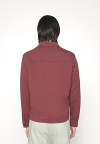 Bordeaux jacka i tjockt tyg, med krage, rygginsyning och knappade ärmslut. Slät struktur utan mönster.