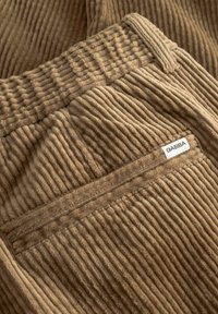 Lichtbruine corduroystof met opvallende verticale ribbels; heeft een zak met een wit "GABBA" label. Zachte textuur en ontspannen ontwerp.