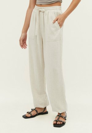 Pantalon beige à larges jambes avec cordon de serrage et grandes poches avant, porté par une personne debout, associé à des sandales plates noires cloutées.