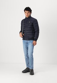 Marineblå pufferjakke med høy krage, glidelås og ribbede mansjetter; har en liten gul logo på brystet, kombinert med lyseblå jeans.