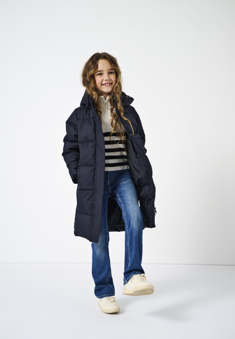 Niño con cabello largo y ondulado que lleva un abrigo acolchado largo negro, suéter a rayas, vaqueros azules y zapatillas blancas, sonriendo con las manos en los bolsillos.