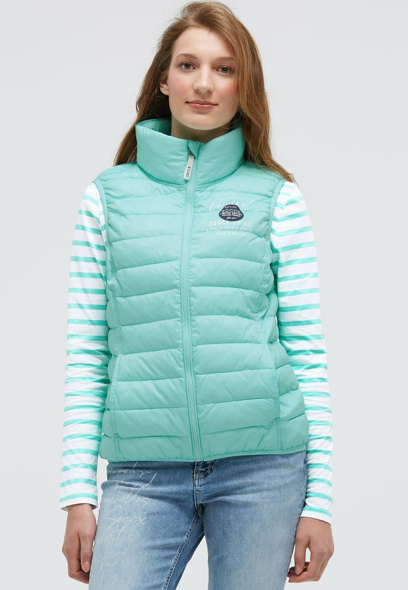 Helles Aqua-puffweste mit hohem Kragen und Reißverschluss, ausgestattet mit einem Logo und Nähedetails. Getragen über einem gestreiften Langarmoberteil.