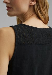 Kvinna med ljus hy och blont hår som bär en ärmlös svart topp med ett spetstajtat övre bakpanel.