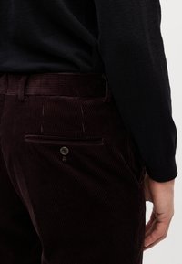 Personne portant un pantalon en velours côtelé marron foncé avec une poche arrière boutonnée et un haut à manches longues noir, la main reposant le long du corps.