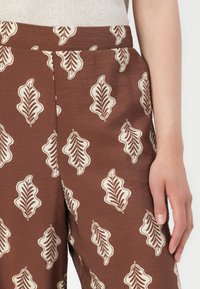 Brune mønstrede bukser lavet af blødt stof med et bladmotiv i creme. Designet inkluderer en sidelomme og en afslappet pasform.