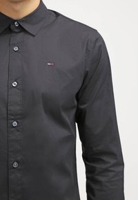 Camisa negra de manga larga con cuello, botones y un pequeño logotipo bordado. Presenta botones brillantes y una textura suave.