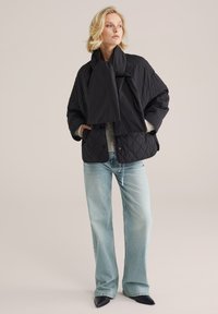 Juodas pūkuotas švarkas su dideliu siluetu, turintis aukštą apykaklę ir pritvirtintą šalį. Nešiojamas virš šviesiai pilko megztinio ir mėlynų džinsų.