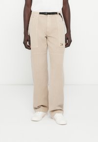 Bézs színű, széles szárú cargo nadrág texturált felülettel, oldalzsebekkel és fekete övvel, fehér tornacipővel párosítva.